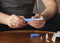 Una persona sostiene un dispositivo de inyección azul en sus manos, con un cartucho adicional y varias agujas desechables sobre una mesa de madera.