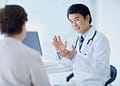 Un médico de cabello oscuro, vestido con una bata blanca y un estetoscopio alrededor de su cuello, está conversando animadamente con una paciente en un entorno moderno de consulta médica, con una computadora y ventanas iluminadas al fondo.