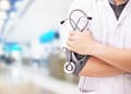 Un profesional de la salud con una bata blanca sostiene un estetoscopio en sus brazos cruzados, con un fondo borroso de un establecimiento médico.