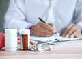 Un médico en bata blanca escribe en una hoja mientras un fonendoscopio y frascos de medicamentos están visibles en la mesa.