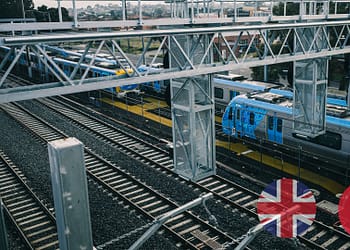 Vista aérea de una estación de tren con vías y trenes azules estacionados, rodeados de un entorno urbano y estructuras metálicas de soporte.
