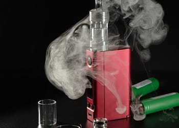 A red vaping device emitting vapor, with two green batteries in the background and a glass cylinder next to it.
