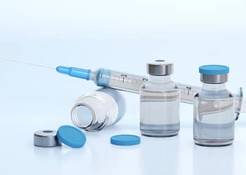 Clear medicine vials with blue caps and a syringe on a light background, some vials are tilted while others are upright.