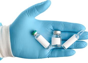 Blue gloved hand holding three vaccine vials, including one with a green cap, one with a silver cap, and a small clear one.
