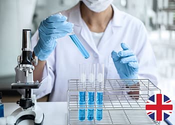 A person in a lab coat and blue gloves holds a test tube with blue liquid, while there are other test tubes in a holder and a microscope on the table.