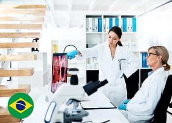 Two female scientists in a laboratory discussing in front of a screen showing microscopic images. One of them is pointing at the screen while the other is watching attentively. On the table there is a microscope and documents.
