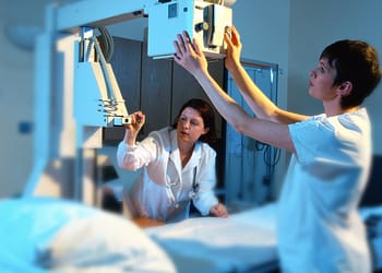 Two healthcare professionals in a medical setting, adjusting X-ray equipment, while one of them watches closely.