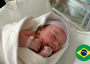 Bebé recién nacido envuelto en una manta blanca, con una pulsera de identificación en la muñeca, mirando hacia la cámara con una expresión de calma. En la esquina inferior derecha, se encuentra una pequeña bandera de Brasil.