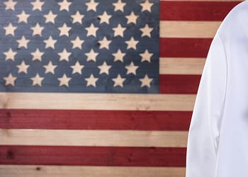 A stethoscope rests on a white coat, in front of a background of the American flag.
