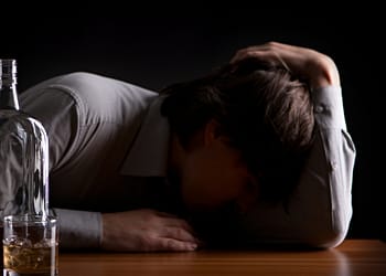 Man in a grey shirt leaning on a wooden table, head down, next to a bottle of liquor and a glass with a drink. Dark background.