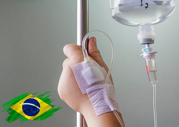 Patient's hand with a drip connected to a stand, in a clinical setting. Blurred background.
