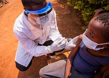 A health care professional wearing protective gear administering a vaccine to a child sitting in a chair outdoors.