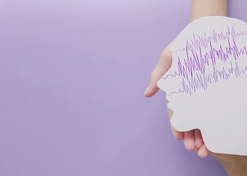 A hand holds a paper cut in the shape of a human profile, with purple lines simulating waves at the top, on a lilac background.