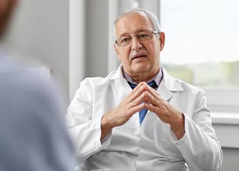 Un médico de mediana edad, con gafas y un bata blanca, está sentado frente a un paciente mientras habla y hace gestos con las manos en una consulta médica iluminada y moderna.