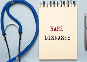 A notebook with the phrase "RARE DISEASES" written in large letters, accompanied by a blue stethoscope and a silver pen on a light background.