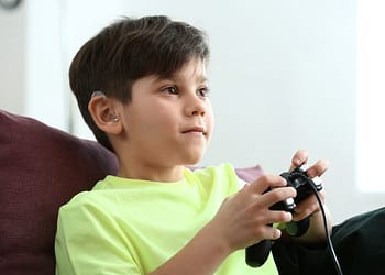 A boy sitting on a couch, using a video game controller and wearing a bright yellow t-shirt, with an earpiece visible in his left ear, displaying a focused expression as he plays the game.