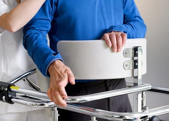 A caregiver holds a person wearing a blue shirt while the person holds onto a harness on a mobility assist device.