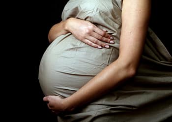 Belly of a pregnant woman covered with a light fabric, with hands resting on her belly, on a dark background.