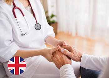 A healthcare professional in a white coat holds the hands of an older patient, showing a gesture of care and empathy in a well-lit, warm indoor setting.