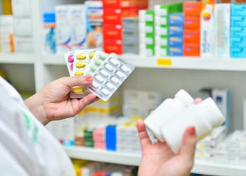 A person holds three blister packs of colored pills and two white bottles of medication, in a pharmacy setting with shelves full of boxes of different colored medications in the background.