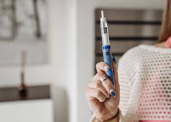A person holds a blue insulin pen in an indoor setting, with minimalist decor in the background and a hand adorned with a ring and painted nails.