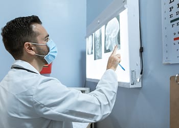 A doctor in a white coat and face mask is pointing at X-ray images under an examination light in a medical office.