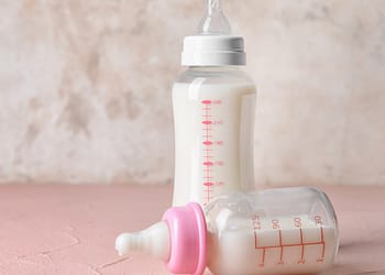 Plastic milk bottles, one white with a white cap and red scale, and one transparent with a pink cap and red scale, on a pink surface.