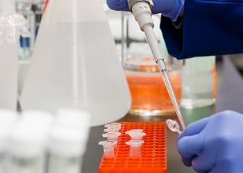 A laboratory with a person using a micropipette to take liquid from a flask, surrounded by test tubes and other scientific tools.