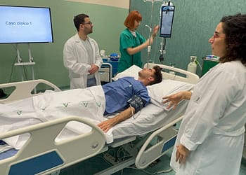 Two doctors and a nurse interact with a patient in a hospital bed. A monitor displays "Clinical Case 1." The patient is semi-reclined with a blood pressure cuff.