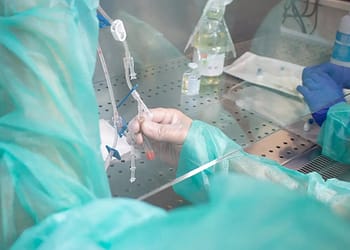 A hand in a clinical setting holds several tubes and syringes, surrounded by laboratory equipment and personal protective equipment, with a background of metallic surfaces and visible pharmaceuticals.