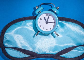 Blue alarm clock on a light blue sleeping mask, on a blue background.