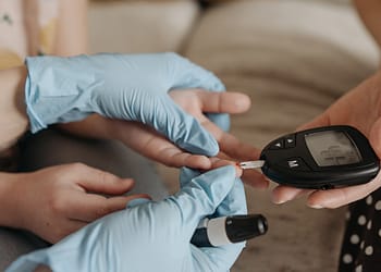 A person wearing latex gloves is using a blood glucose monitoring device on another hand, while preparing to apply a blood sample.