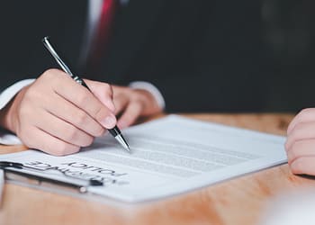 A hand writes with a pen on a document on a table, while another hand rests next to the document. The visible text includes the phrase "Policy."