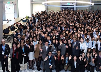 Large group of people of diverse ages and backgrounds, smiling and waving at the camera at an event, with a festive atmosphere in a large indoor space.