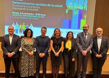 A group of eight people posing in front of a poster advertising an event on social determinants of health at La Paz University Hospital, Madrid.