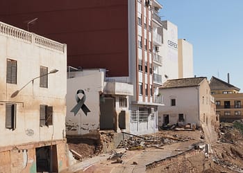 Image of a damaged urban area with crumbling buildings and visible debris; a symbolic black ribbon can be seen on the left side.