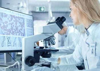 Laboratory woman looking through a microscope, with a computer screen showing scientific graphs and data.