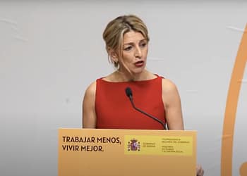 Woman with blonde hair and red dress, speaking from a podium with a sign that says "WORK LESS, LIVE BETTER."