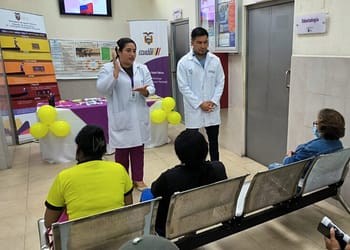 Two health professionals, dressed in white coats, are speaking in front of a group of people sitting in a waiting room. There are yellow balloons decorating the table in the background and informative posters on the walls.