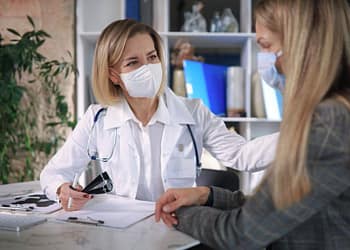 A female doctor wearing a mask, wearing a white coat and a stethoscope, is talking to a patient, also wearing a mask, in an office. The doctor is holding a pencil, and a table with documents and a plant can be seen in the background.