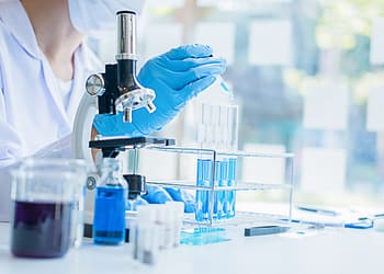 Un científico en un laboratorio utiliza una pipeta para añadir líquido a tubos de ensayo que contienen soluciones azules, mientras un microscopio y otros instrumentos de laboratorio están visibles en la mesa.