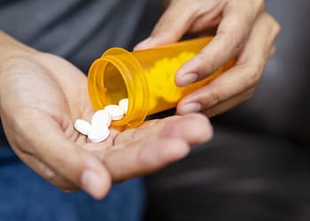 One hand holds an orange bottle of medicine, while another hand receives white pills that fall from the bottle into the palm.