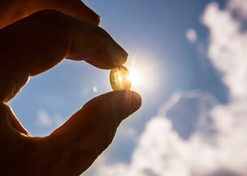 A hand holds a small transparent capsule against a background of blue sky with clouds, while sunlight shines through the capsule, creating a luminous effect.