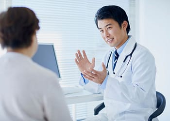 Un médico de cabello oscuro, vestido con una bata blanca y un estetoscopio alrededor de su cuello, está conversando animadamente con una paciente en un entorno moderno de consulta médica, con una computadora y ventanas iluminadas al fondo.
