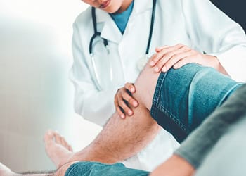 A doctor examines a patient's knee while they are both seated. The doctor is wearing a white coat and has a stethoscope around his neck.