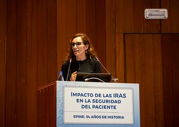 Mujer de cabello rizado y gafas, hablando en un podio con un letrero que dice "Impacto de las IRAS en la seguridad del paciente". Fondo de madera.