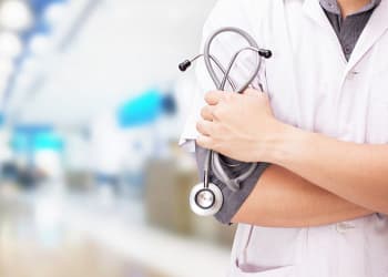 Un profesional de la salud con una bata blanca sostiene un estetoscopio en sus brazos cruzados, con un fondo borroso de un establecimiento médico.