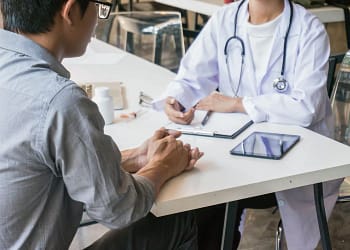 Un hombre sentado frente a un médico que lleva una bata blanca y un estetoscopio, en un consultorio con una mesa, una tableta y varios frascos de medicamentos visibles. El médico parece estar explicando algo al paciente.