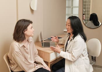 Una mujer con cabello castaño sentada, escuchando atentamente a una doctora de piel oscura con una bata blanca, que está hablando animadamente frente a una mesa con una laptop, en un consultorio moderno con paredes beige y un espejo circular en la pared.