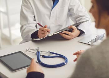 Un médico, con una bata blanca y un bolígrafo en la mano, examina un documento mientras escucha a una paciente sentada frente a él. En la mesa hay un estetoscopio y una tableta.